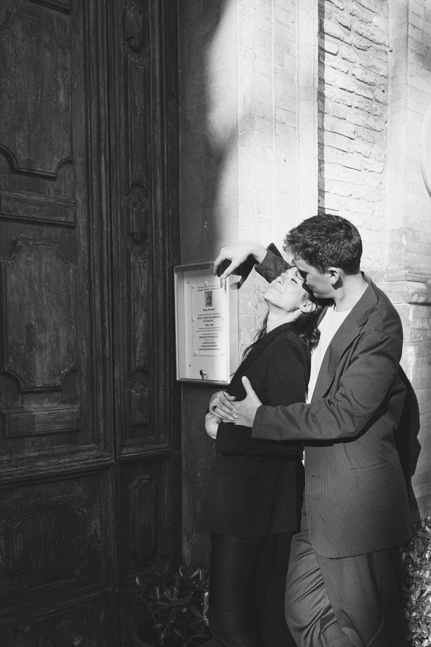 Black and white photo of a couple embracing and kissing by a large wooden door against a stone wall. Italy destination wedding.