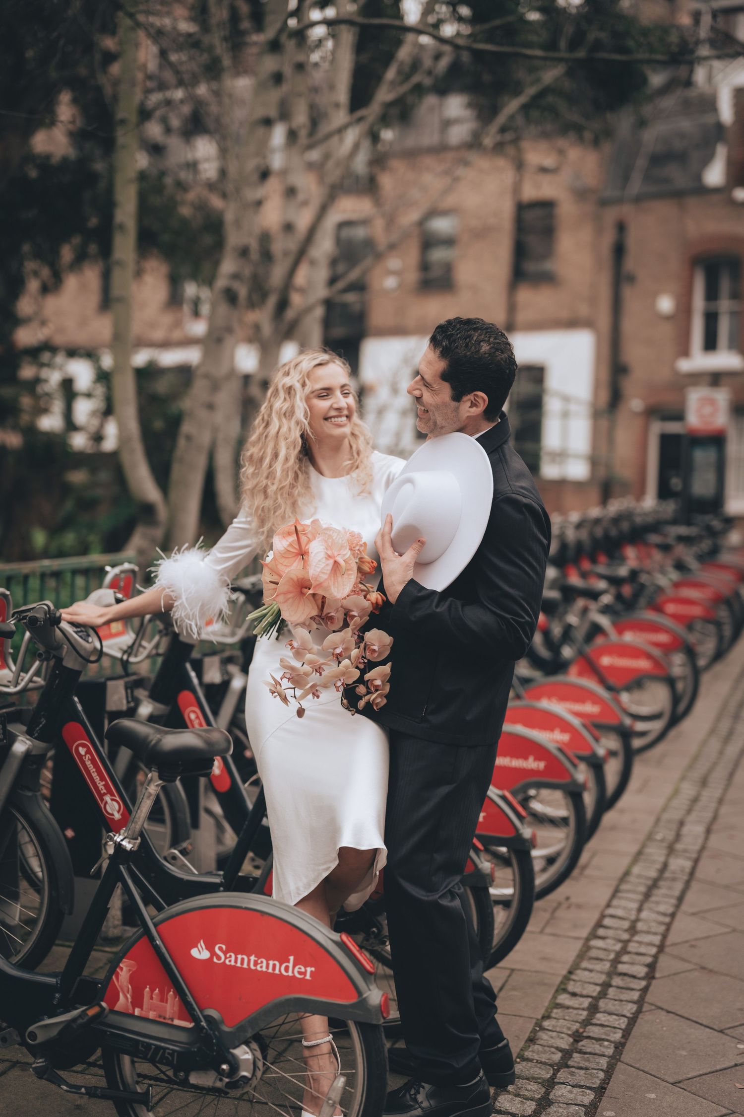 Bride and groom laughing together beside Santander bike hire station on a London street, bride holding floral bouquet. London elopement.