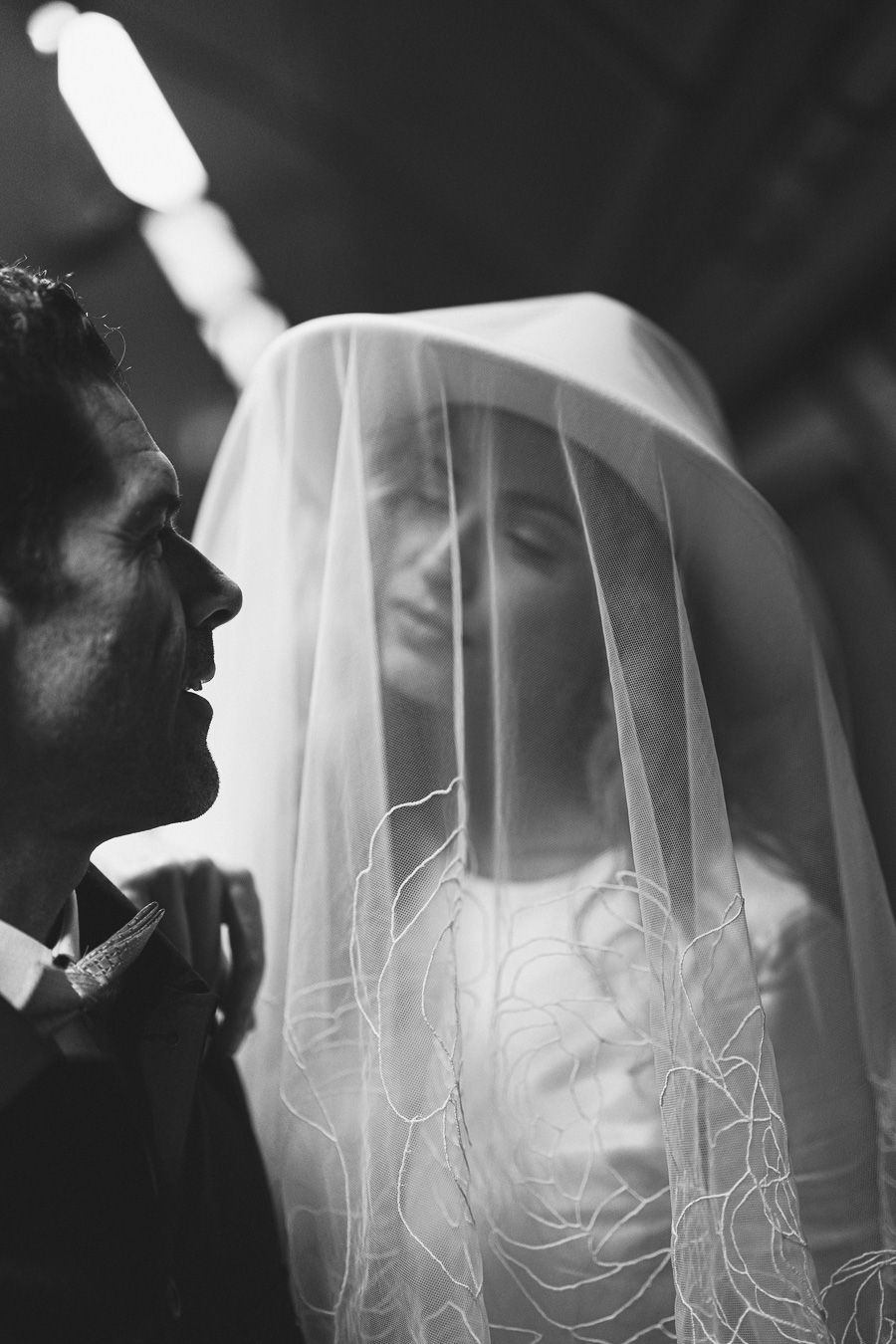 Black and white wedding photo of groom gazing at bride through delicate embroidered veil in romantic portrait in London tube station.