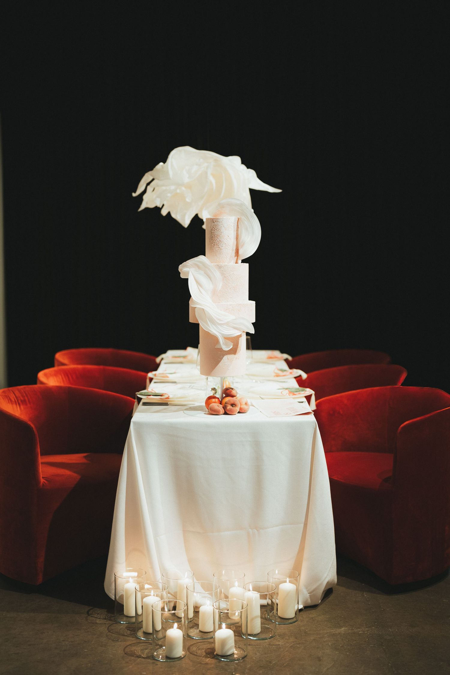 Elegant wedding cake on white draped table surrounded by candles and red velvet chairs against dark background.