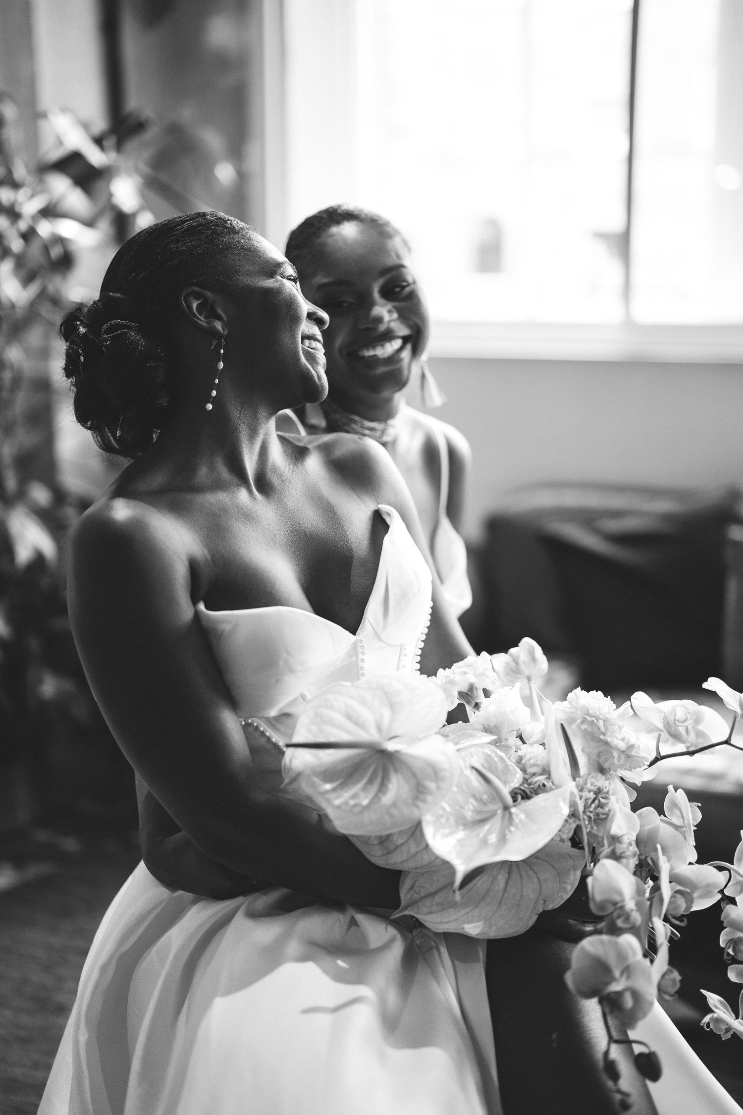 Bride and bridesmaid laugh joyfully, one holding a bouquet of orchids, in a black and white wedding photo.