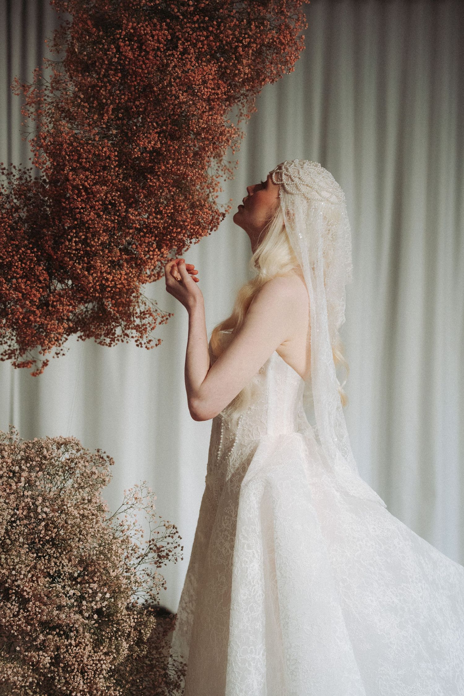 Bride in lace gown and veil reaches toward dried red flowers against soft curtain backdrop.