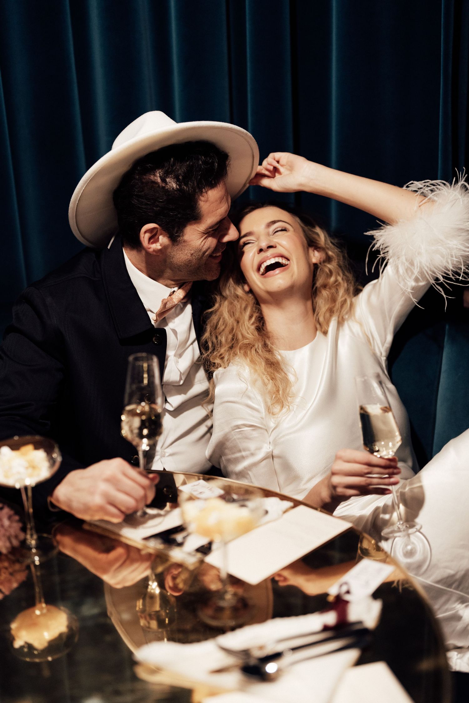 Elegant couple laughing and celebrating with champagne at a formal dinner, woman in white dress, man in suit and hat.