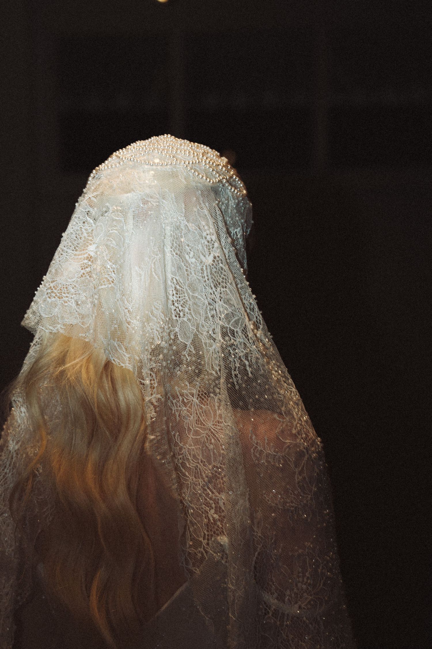 Bride wearing an ornate lace wedding veil with pearl headpiece, long blonde hair visible from behind.