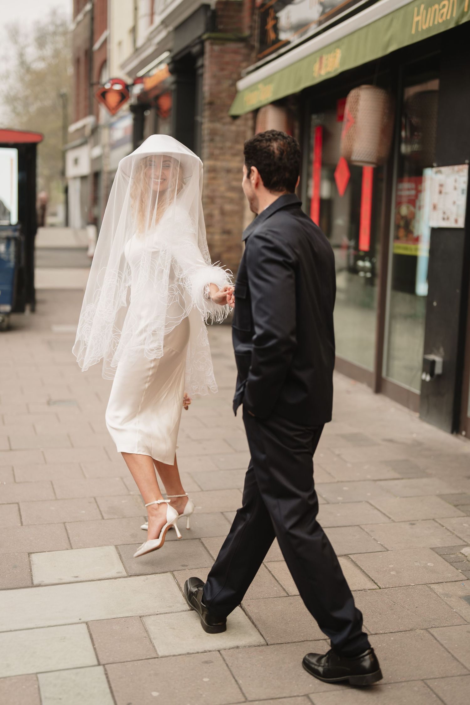 A bride in a white dress and veil walks hand-in-hand with a groom in a black suit on a London city sidewalk. London elopement wedding.