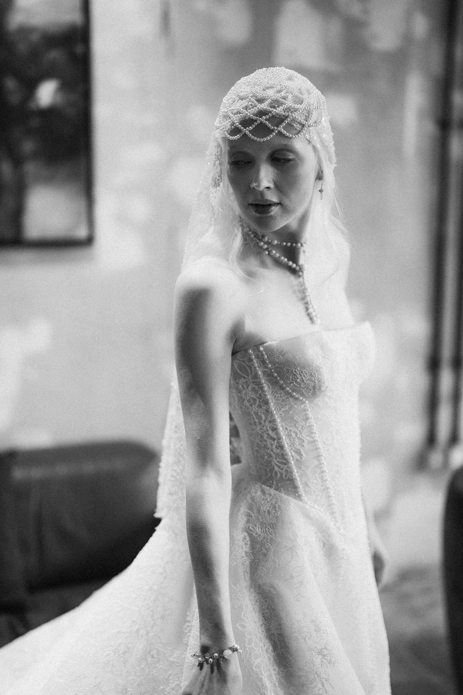 Black and white photo of a bride in lace gown, pearl necklace, and ornate mesh headpiece with veil.