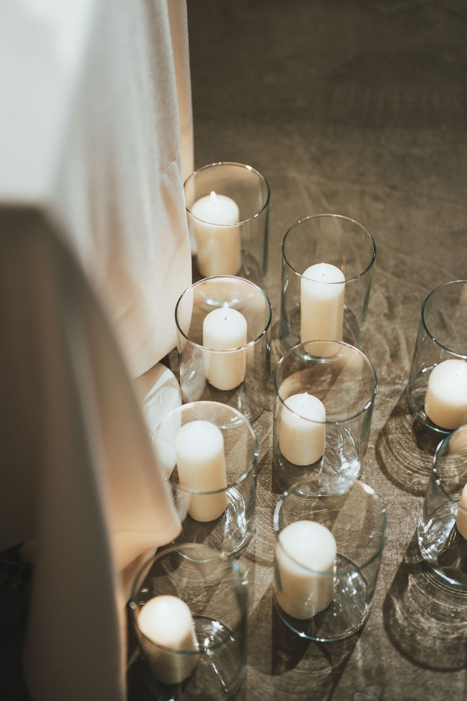 White pillar candles in glass hurricane vases arranged on floor beside draped fabric for elegant event decor.