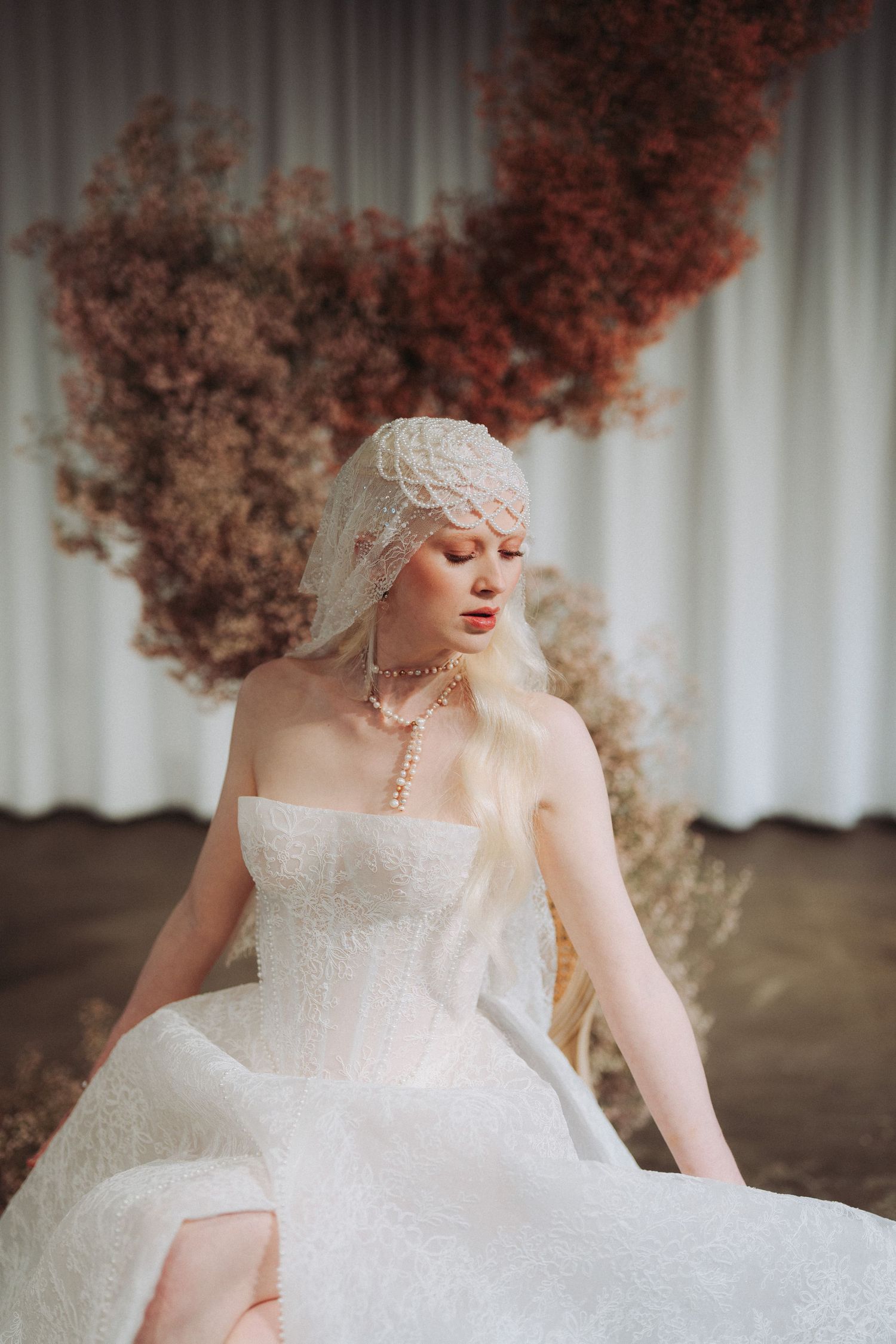 Elegant bride in white lace gown and veil cap, adorned with pearl necklace, posed before dried floral backdrop.