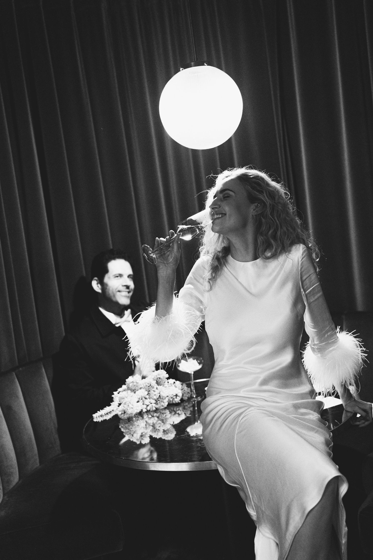 Black and white photo of a smiling bride in white dress sipping champagne beside a groom in a dimly lit venue.