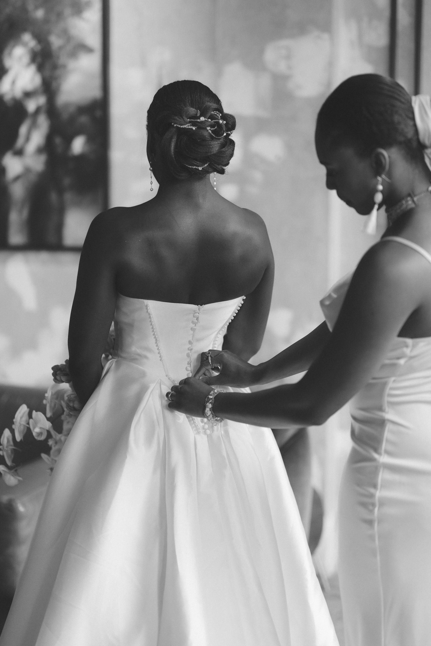 Black and white photo of a bride in a strapless gown being buttoned up by her bridesmaid before the wedding.