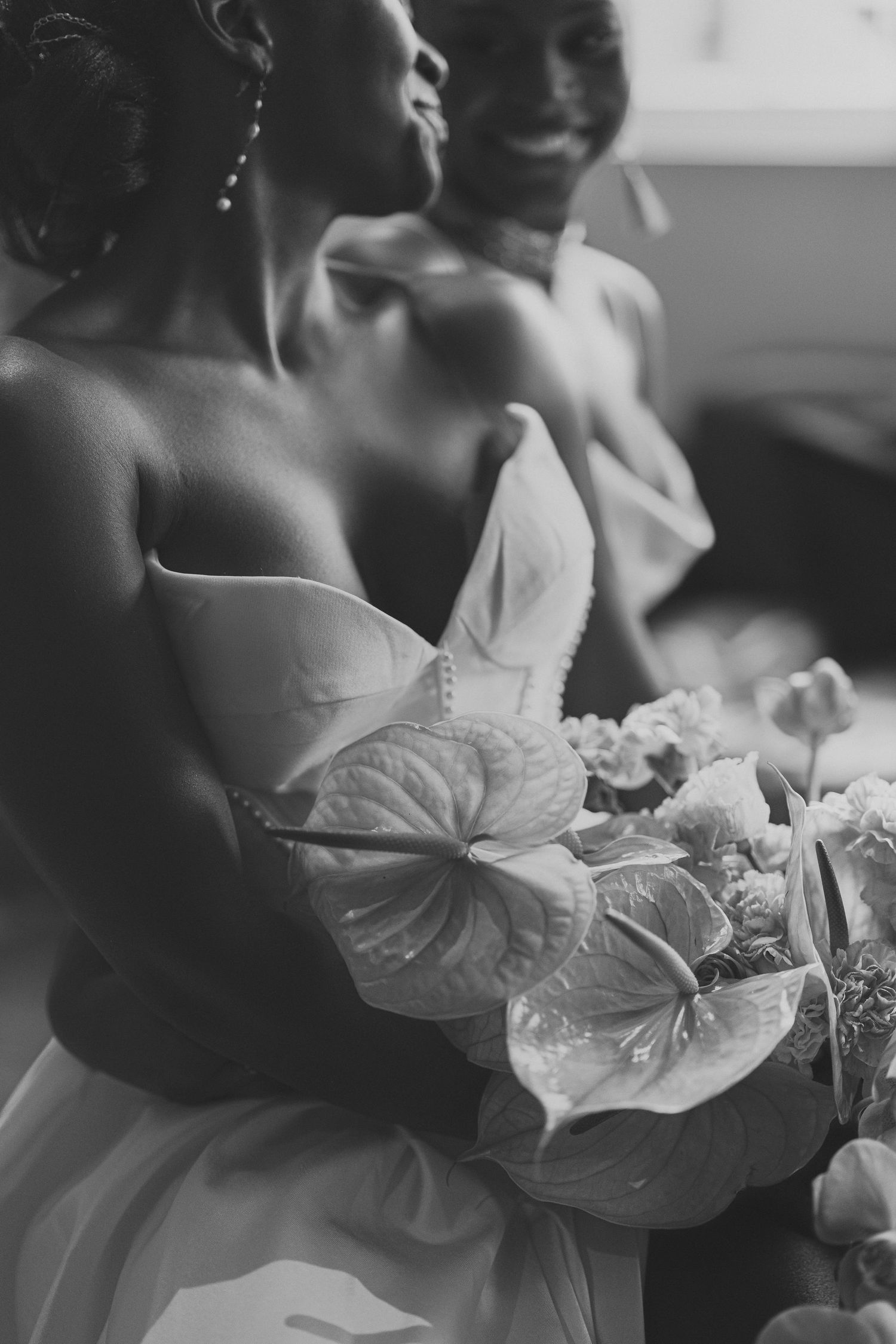 Black and white photo of a bride holding an anthurium bouquet, with a smiling groom in the background.