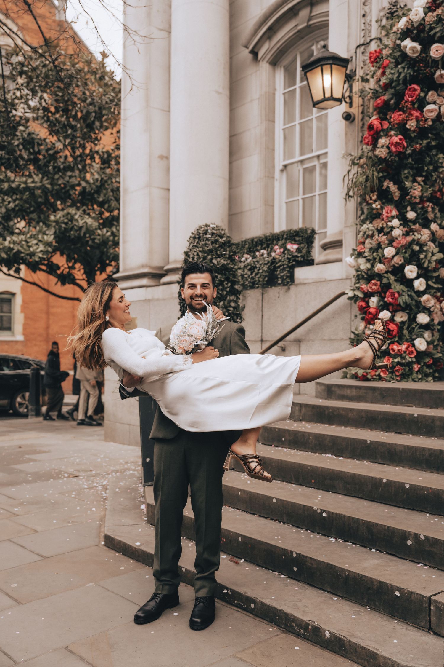 Groom lifting bride in white dress outside flower-adorned Chelsea Old Town Hall steps, confetti falling, joyful wedding celebration.