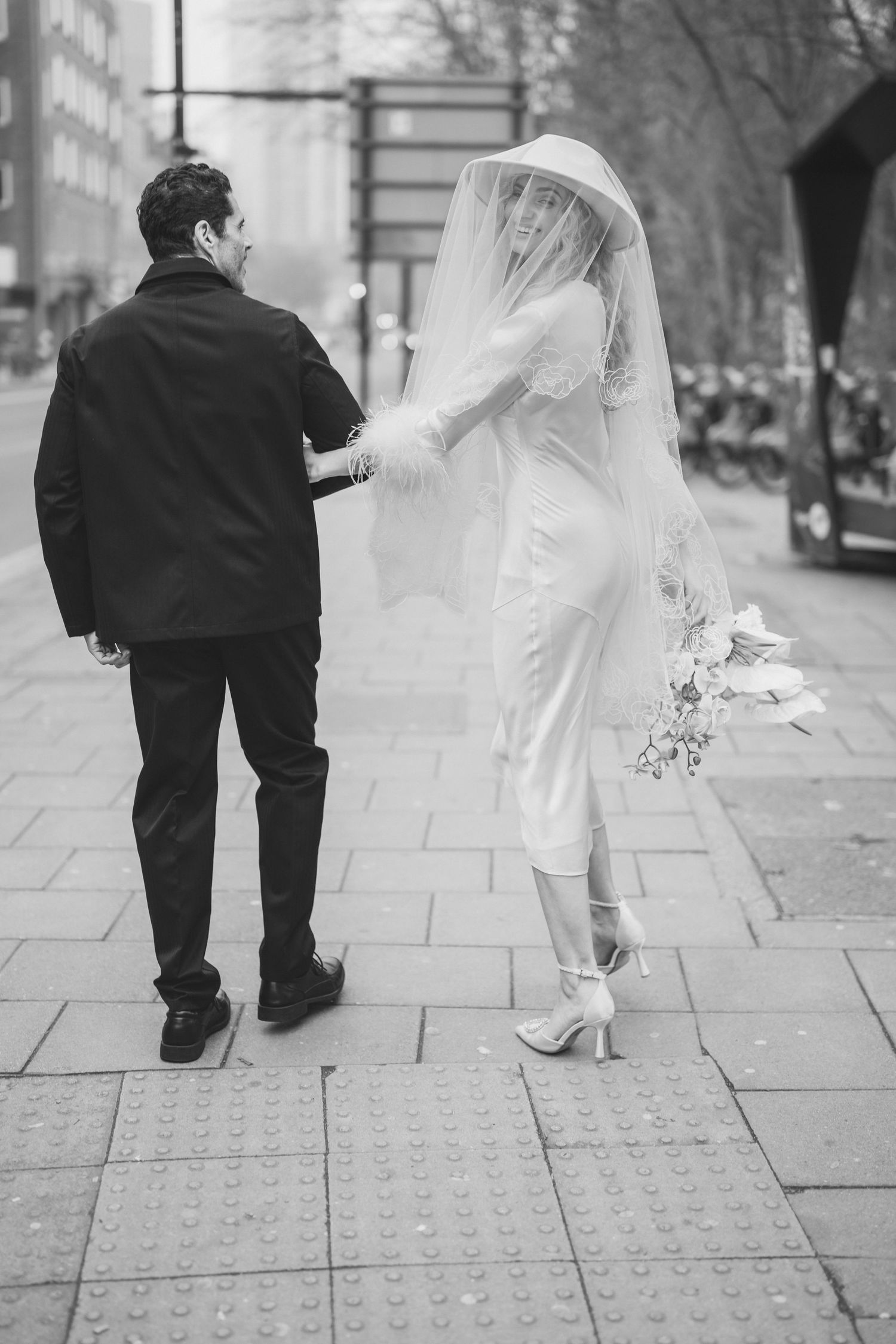Black and white photo of a bride and groom walking hand in hand on a city sidewalk, bride wearing veil and holding bouquet.