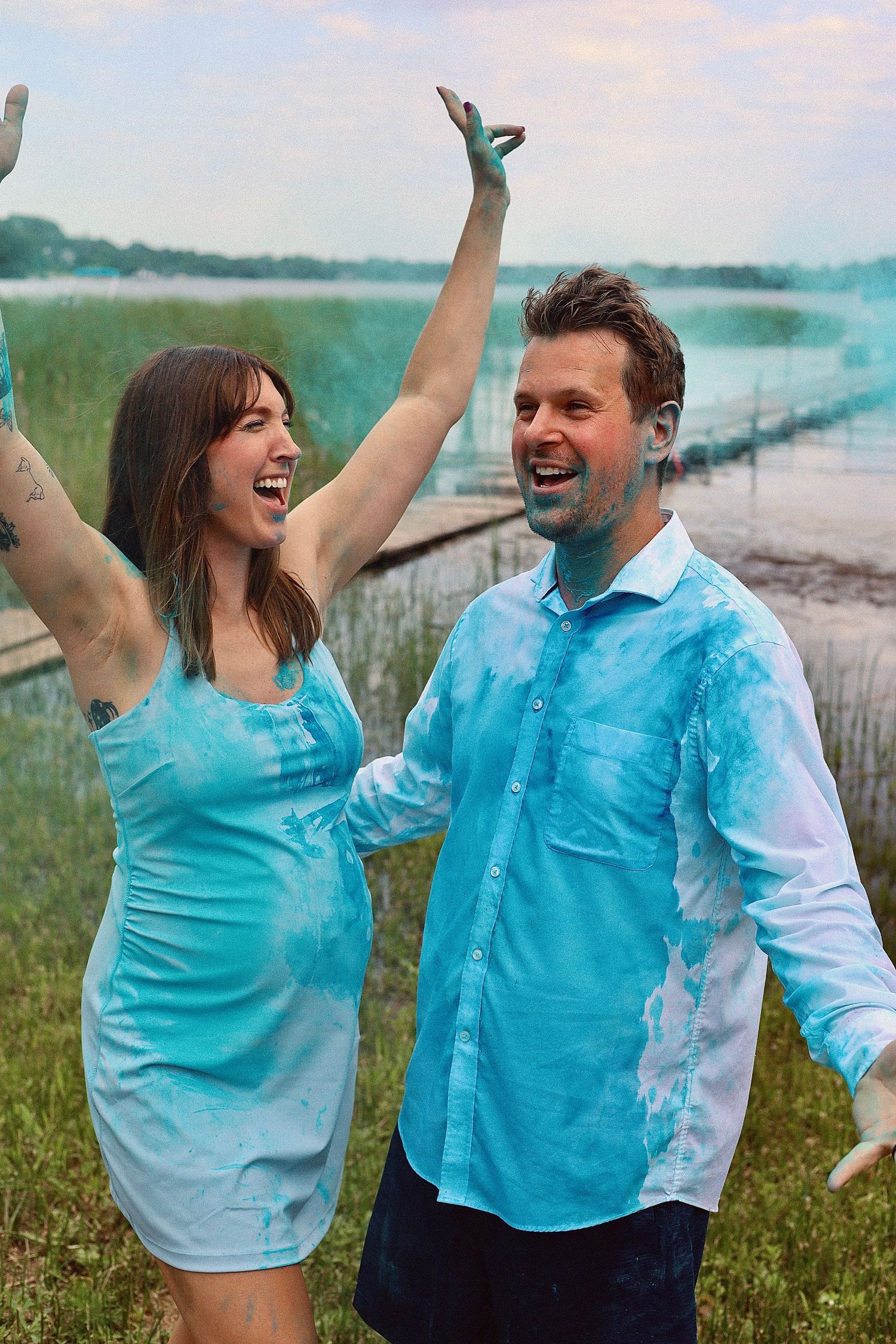 A couple celebrates with joy during a blue powder reveal outdoors by the water.