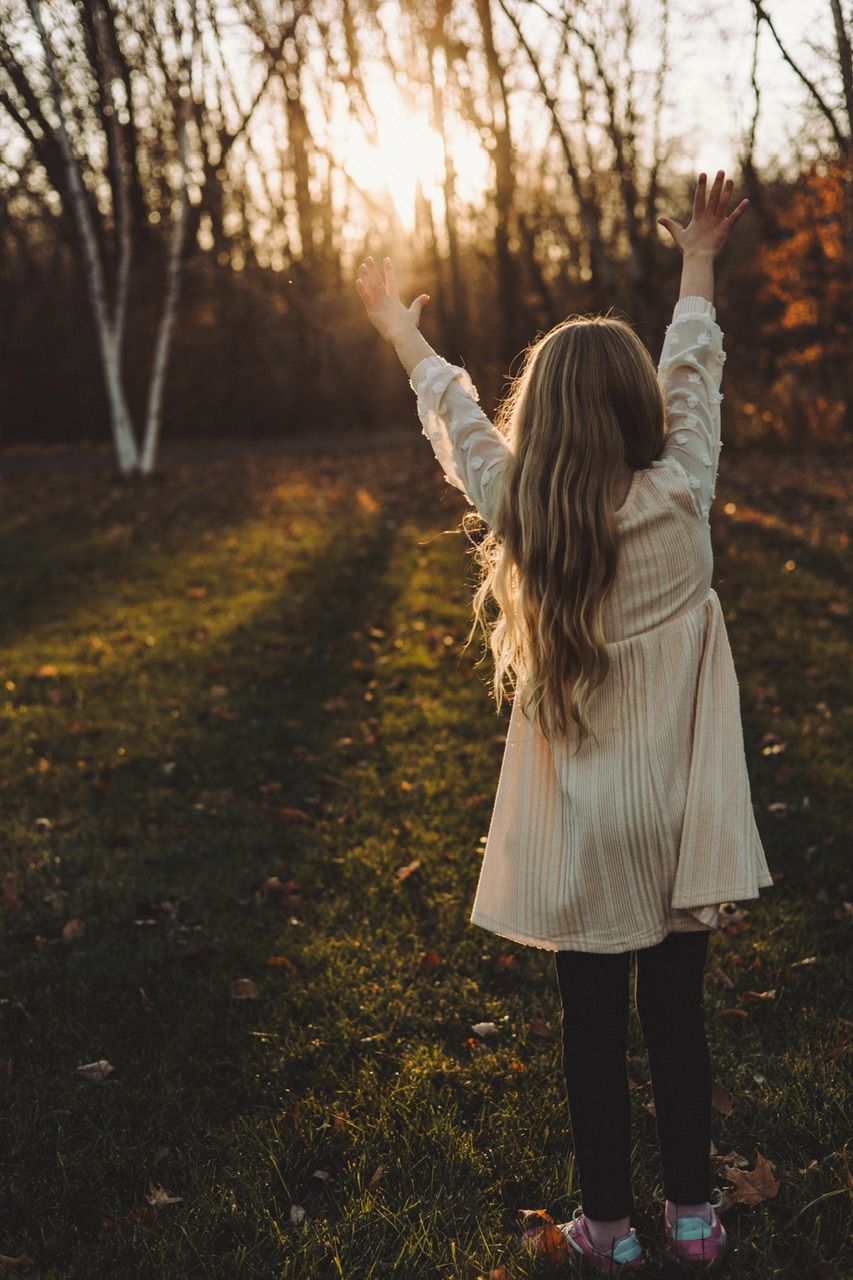 Person in cream sweater raises arms joyfully against sunset in autumn forest.