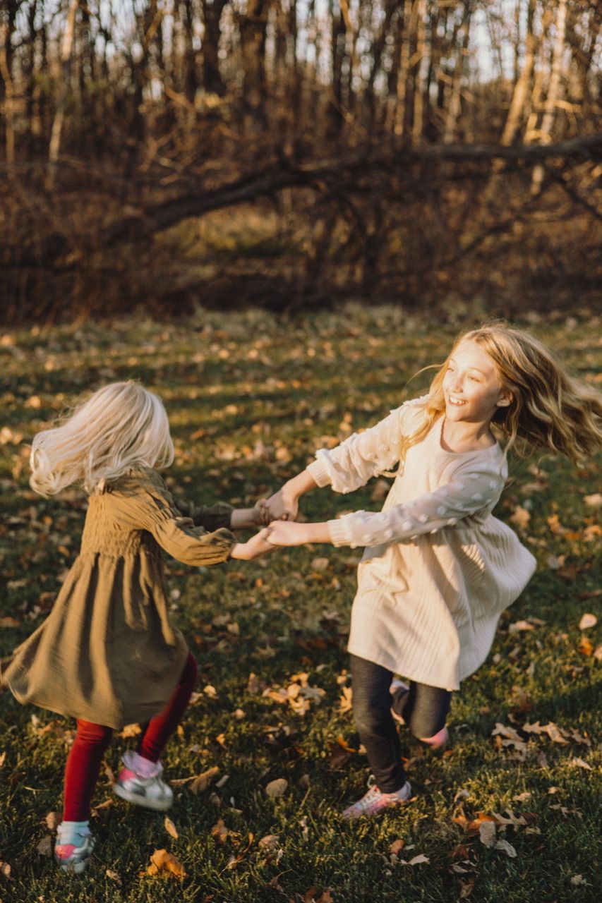 Two children in fall clothing play and twirl together on grass covered with autumn leaves.