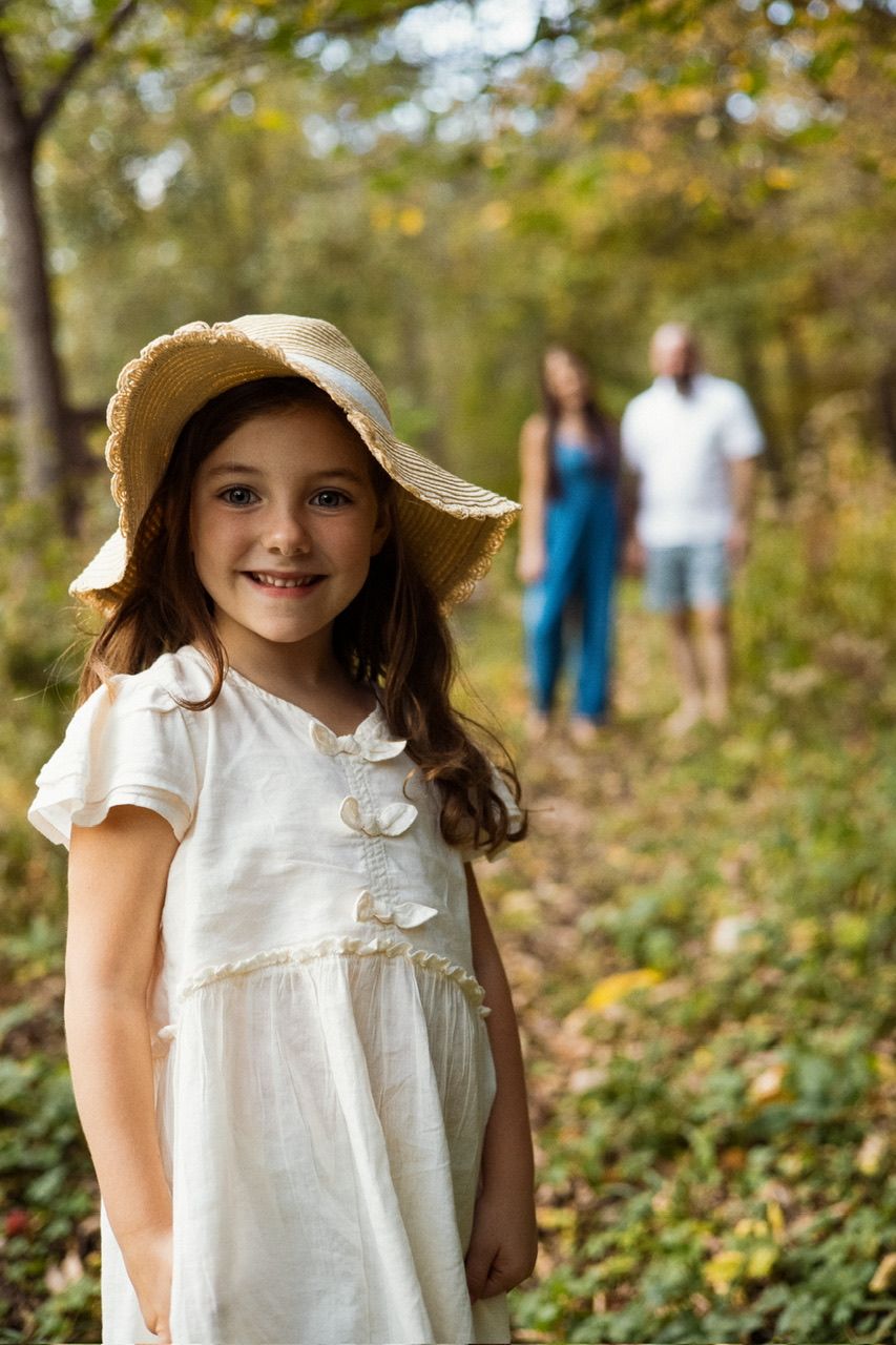 Child in white dress and sunhat poses on forest path with two people visible in background.