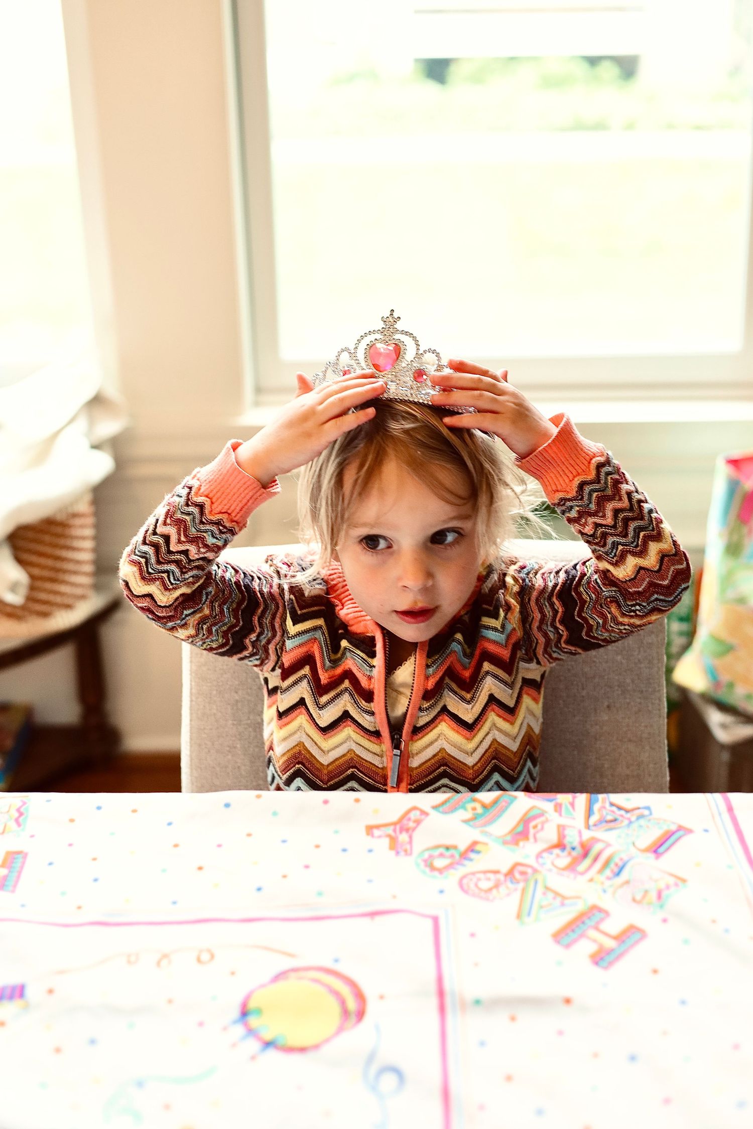 Child adjusting a sparkly tiara while sitting at an art-covered table.