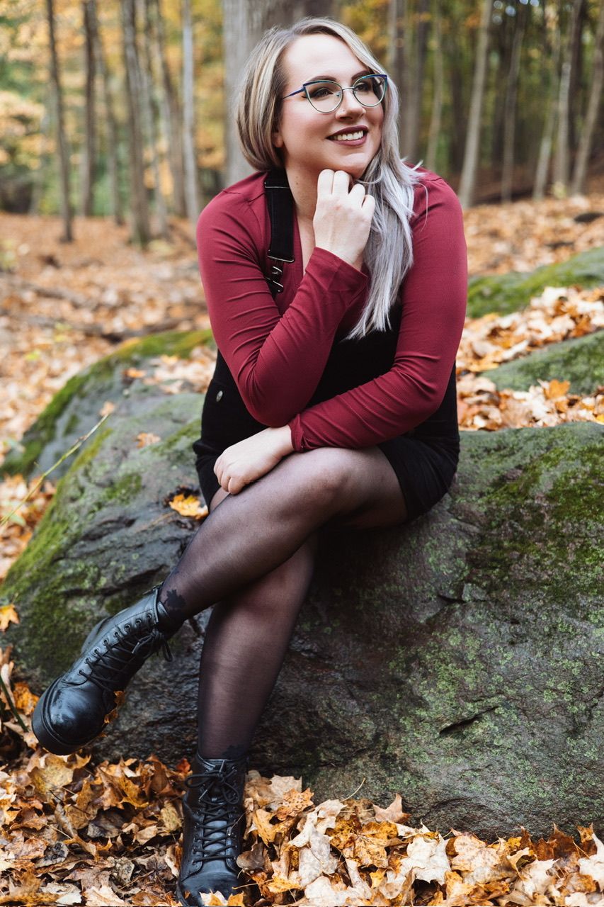 Person in burgundy sweater and tights sits on mossy rock surrounded by fallen autumn leaves.