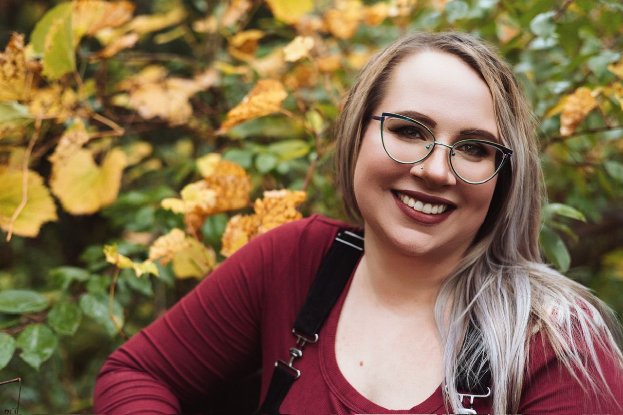 A person wearing a burgundy sweater and glasses poses outdoors with autumn foliage in the background.