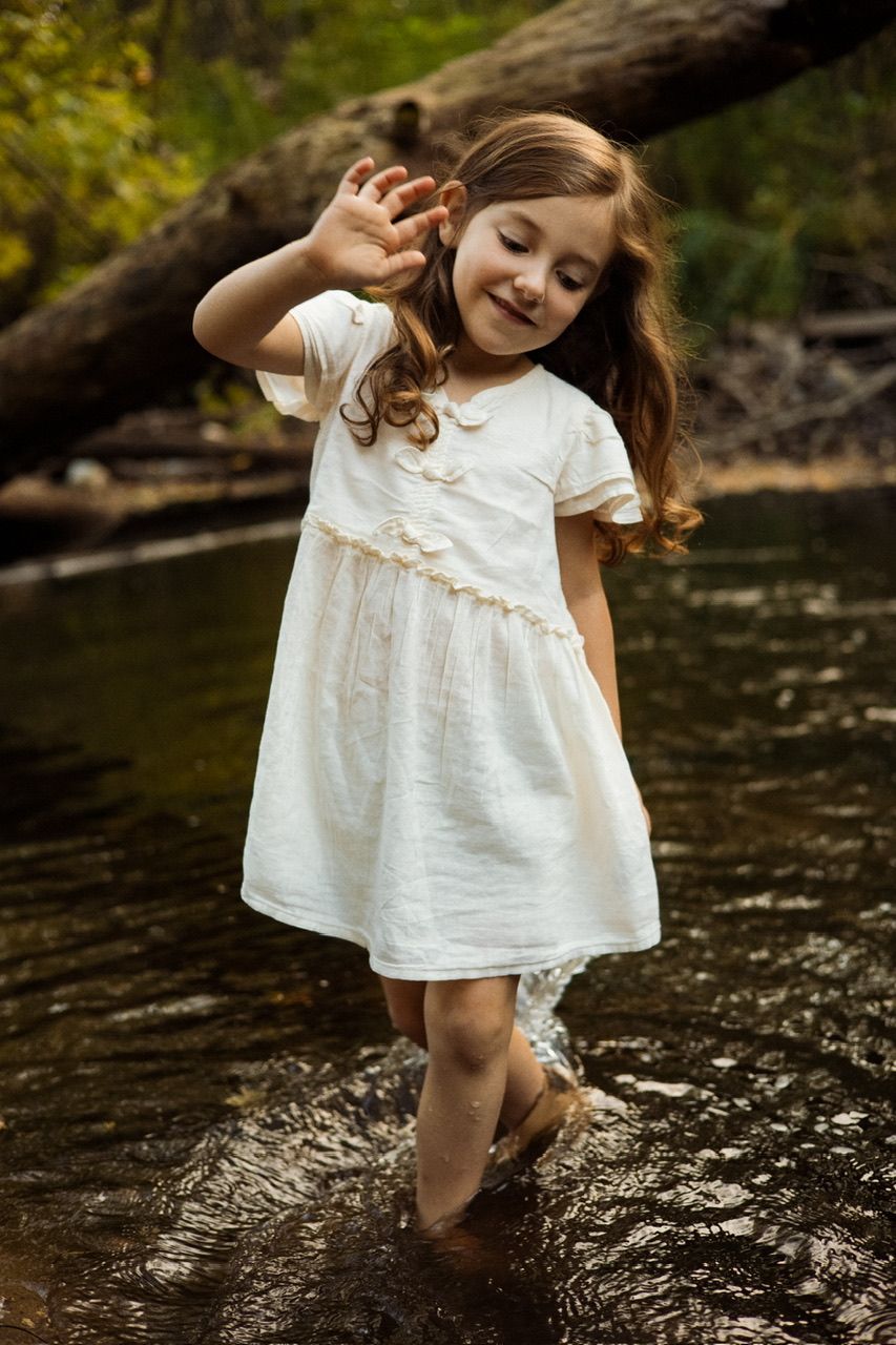Child in white dress playing and splashing in shallow stream water.