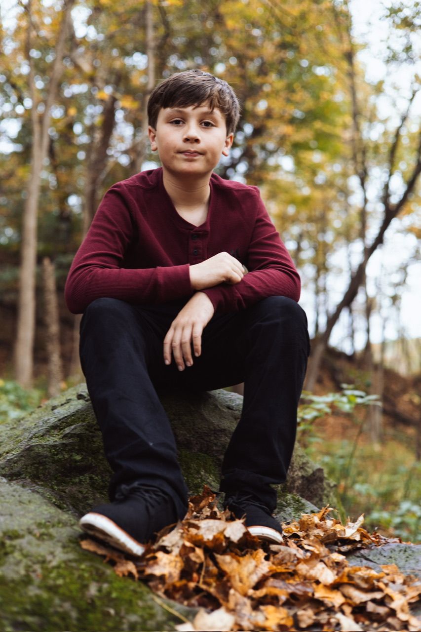 Person in burgundy sweater sits casually on rock surrounded by fallen leaves in wooded setting.