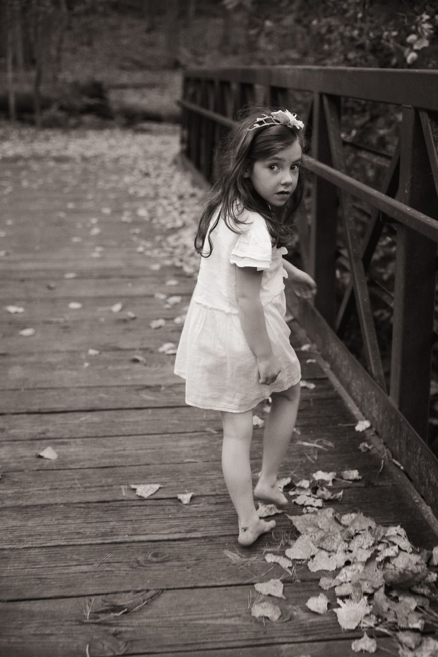 Black and white photo of someone in white dress looking back while standing on wooden bridge with fallen leaves.