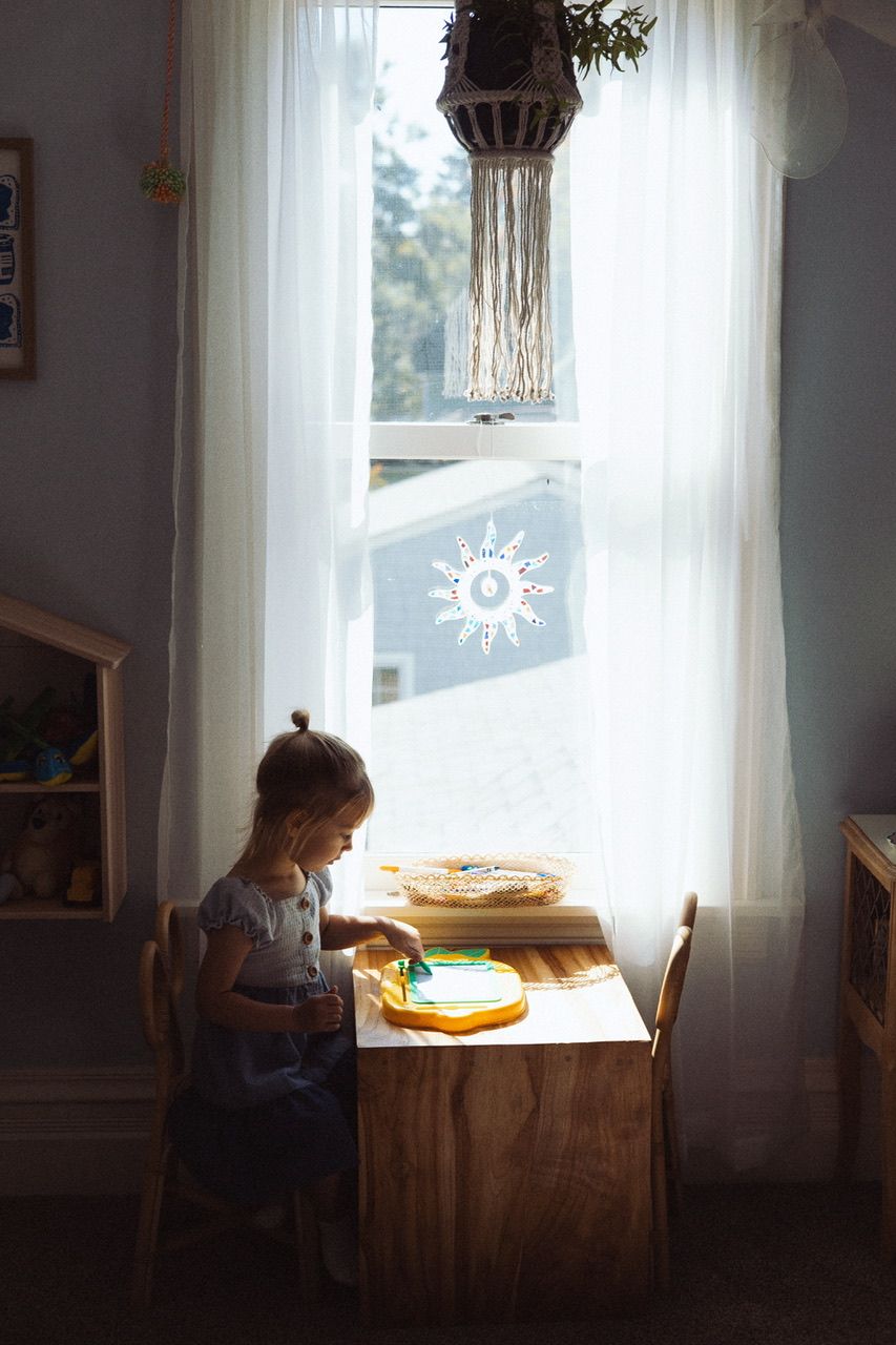 Sunlit room with child at wooden table by window with hanging decorations.