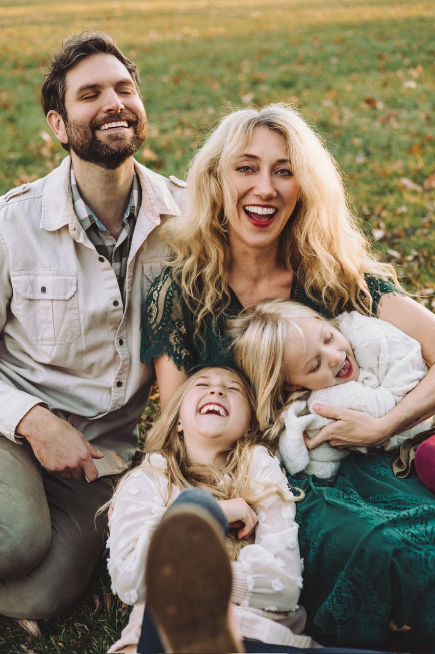 A joyful family laughs together while sitting on grass during an outdoor photo session.