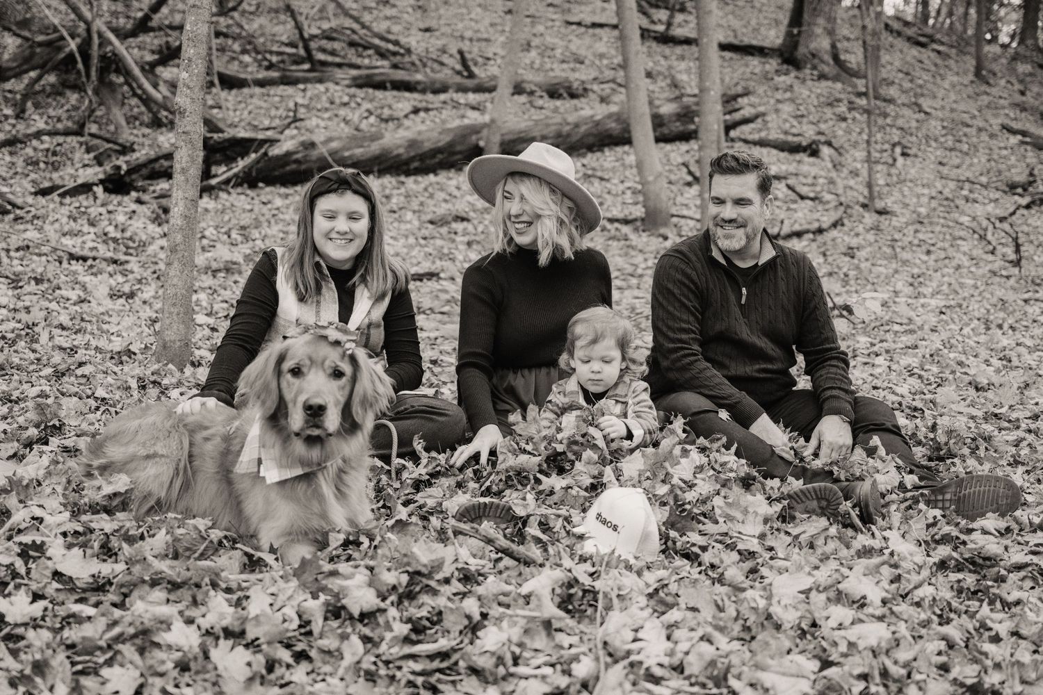 Family and their golden retriever sitting together on fallen autumn leaves in the woods.