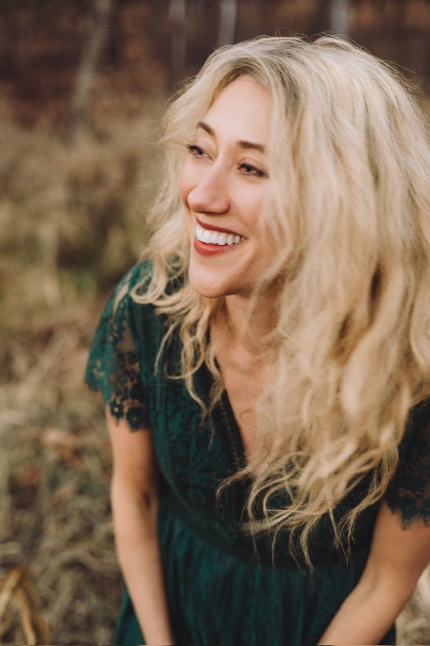 Person with blonde hair wearing a dark green top laughs joyfully in an outdoor autumn setting.