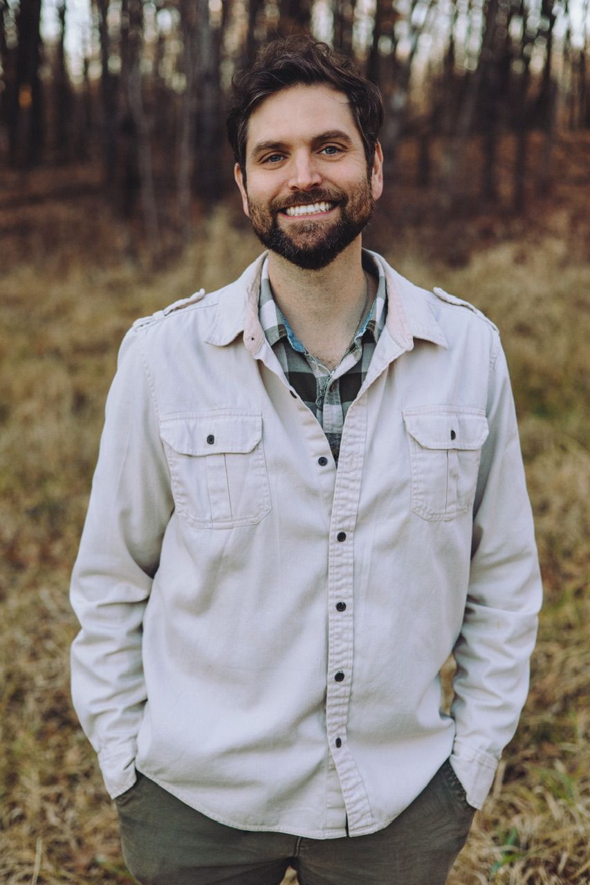 Person wearing a white button-up shirt poses casually outdoors in autumn woodland setting.