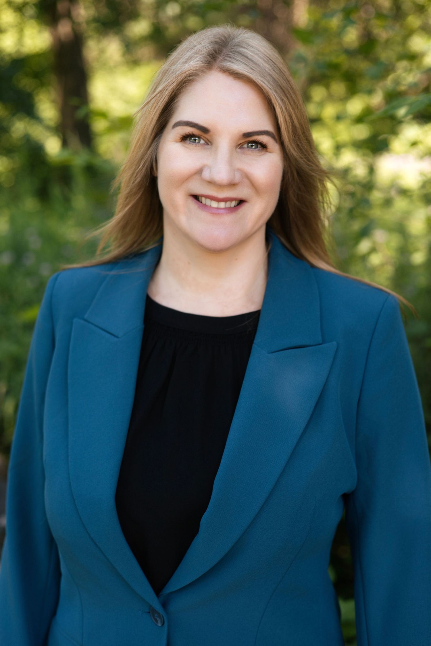 Professional headshot portrait in a navy blazer and black top against a natural outdoor background.