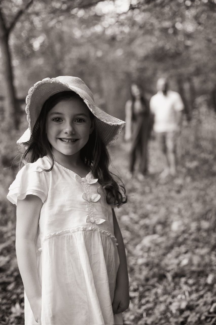 Black and white photo of someone in a white dress and sunhat standing in a wooded area.