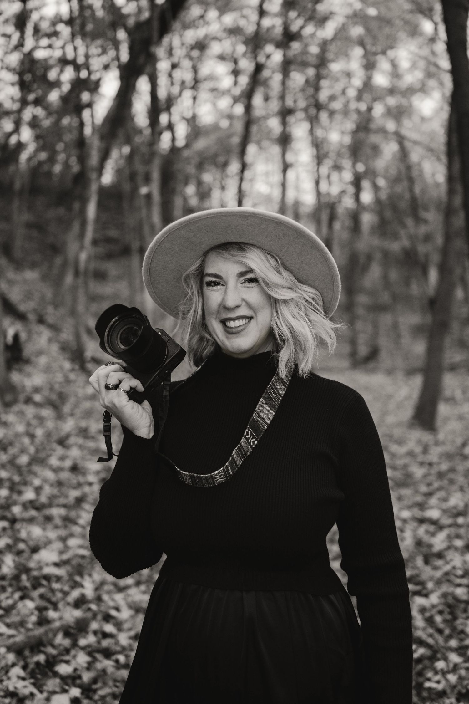 Person in black coat and wide-brimmed hat poses playfully in autumn forest setting.