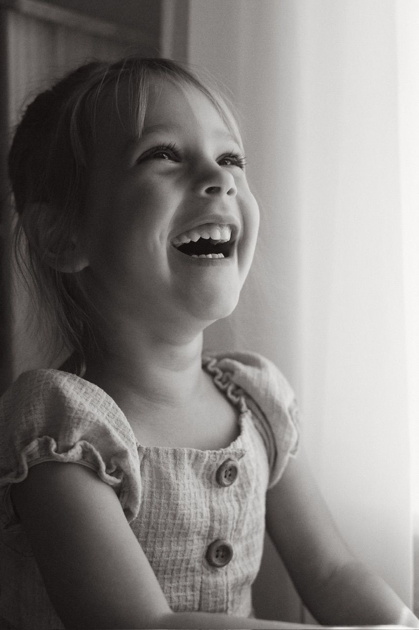 Child in a white dress laughing joyfully by a window.
