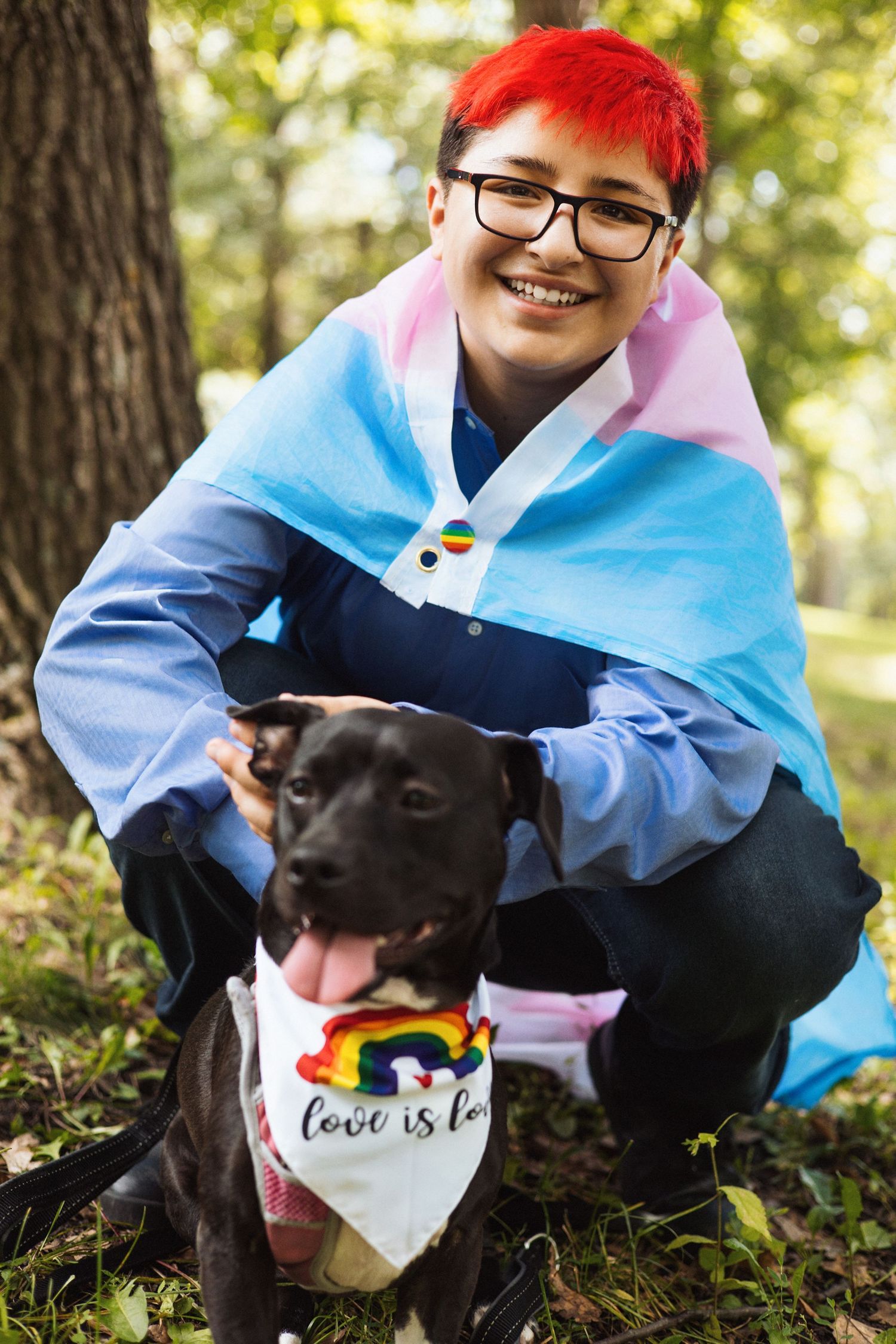Person in blue shirt poses with black dog wearing rainbow bandana in sunny forest setting.