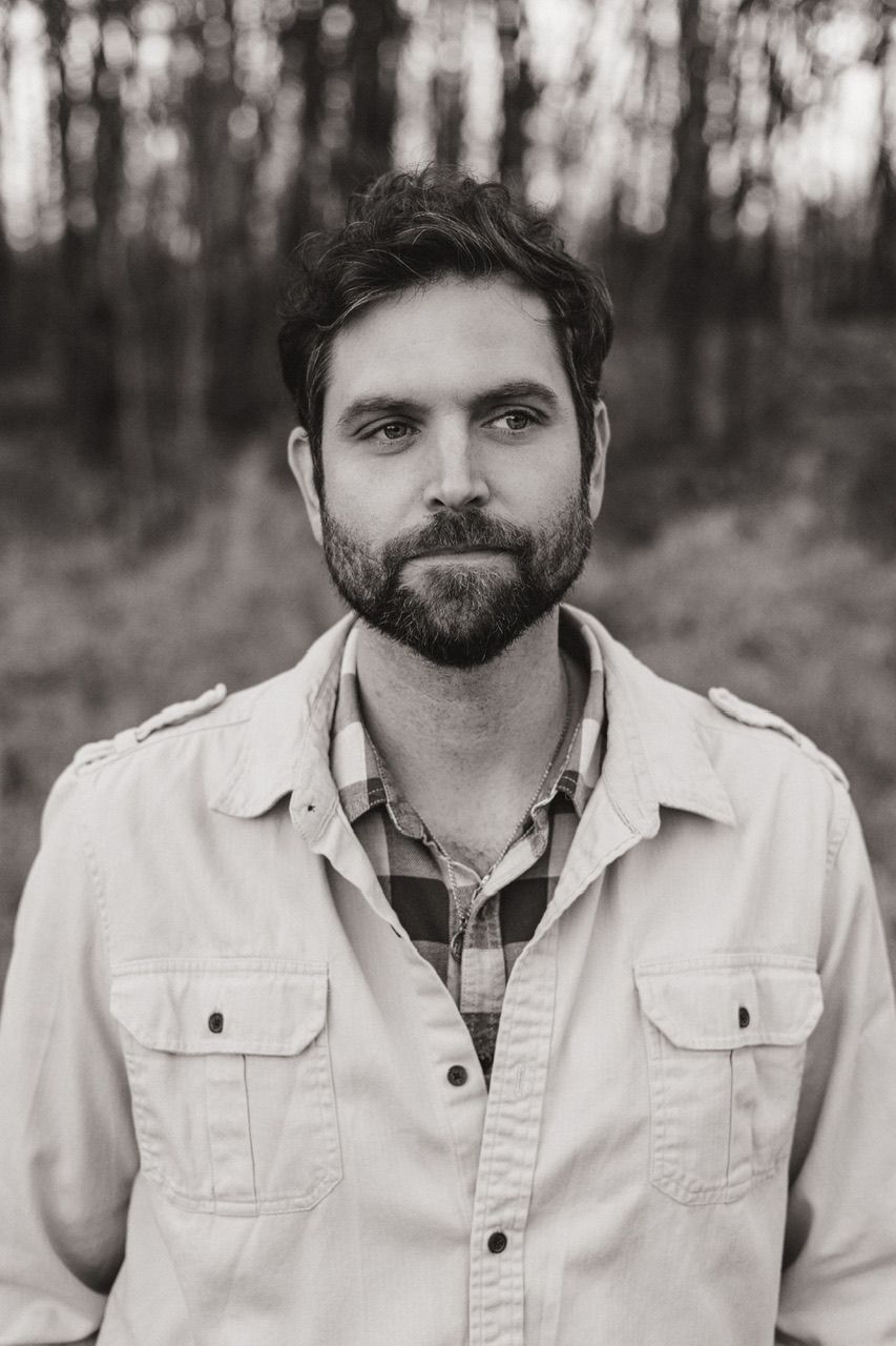 Portrait of a person in a light jacket and plaid shirt against a blurred forest background.