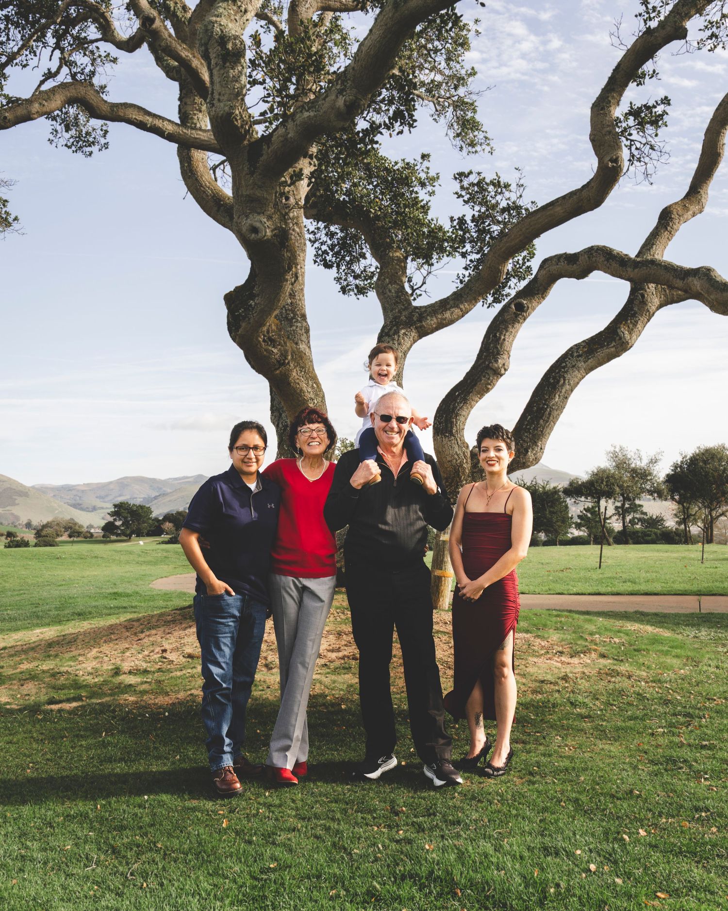 A group of people pose together under a large twisted tree on a grassy hillside with mountains in the distance.