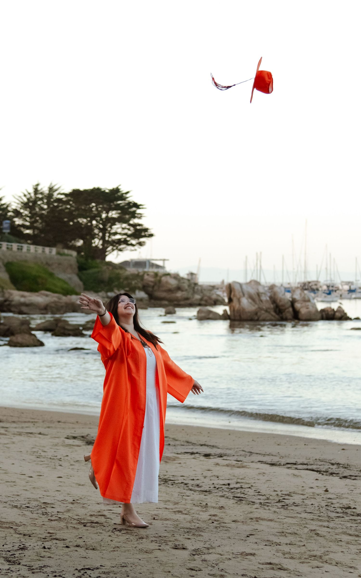 Woman in orange robe tossing a red hat on a sandy beach with rocky shoreline and marina in background.
