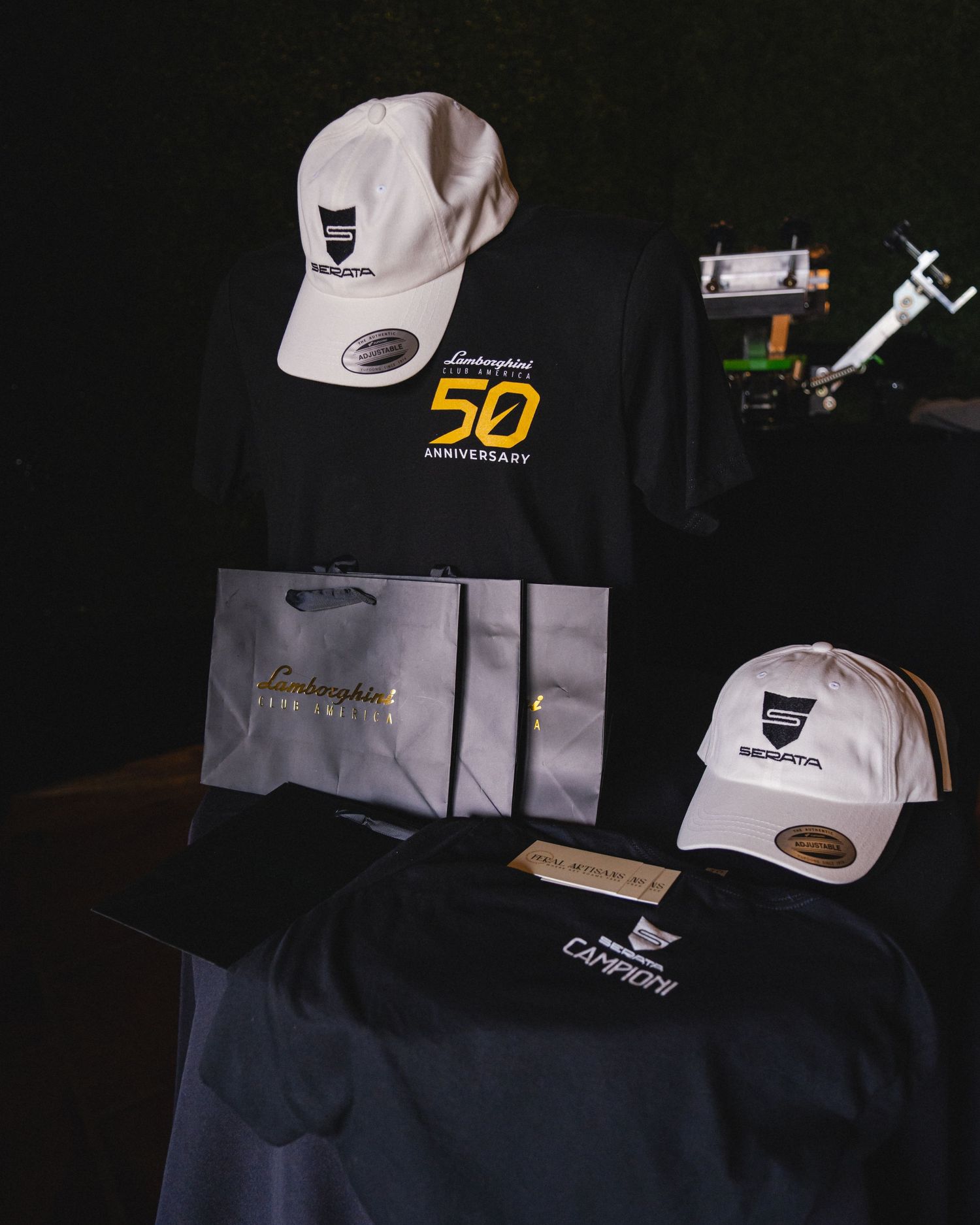 Lamborghini 50th Anniversary merchandise set featuring white caps, black t-shirts, and branded gift bags.