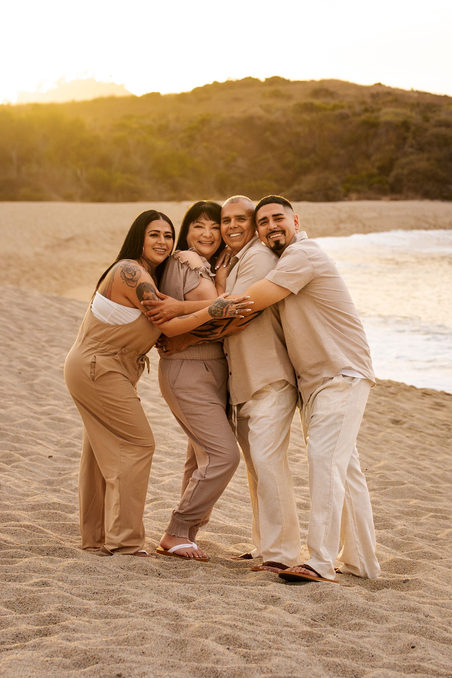 A group of people wearing beige and white outfits pose playfully together on a sandy beach at sunset.