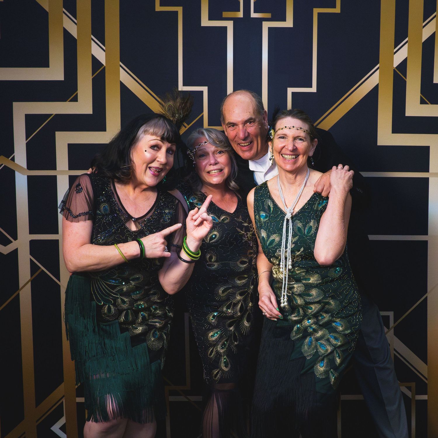 Four adults in green peacock-feathered flapper dresses and 1920s Great Gatsby attire pose at an Art Deco themed party.