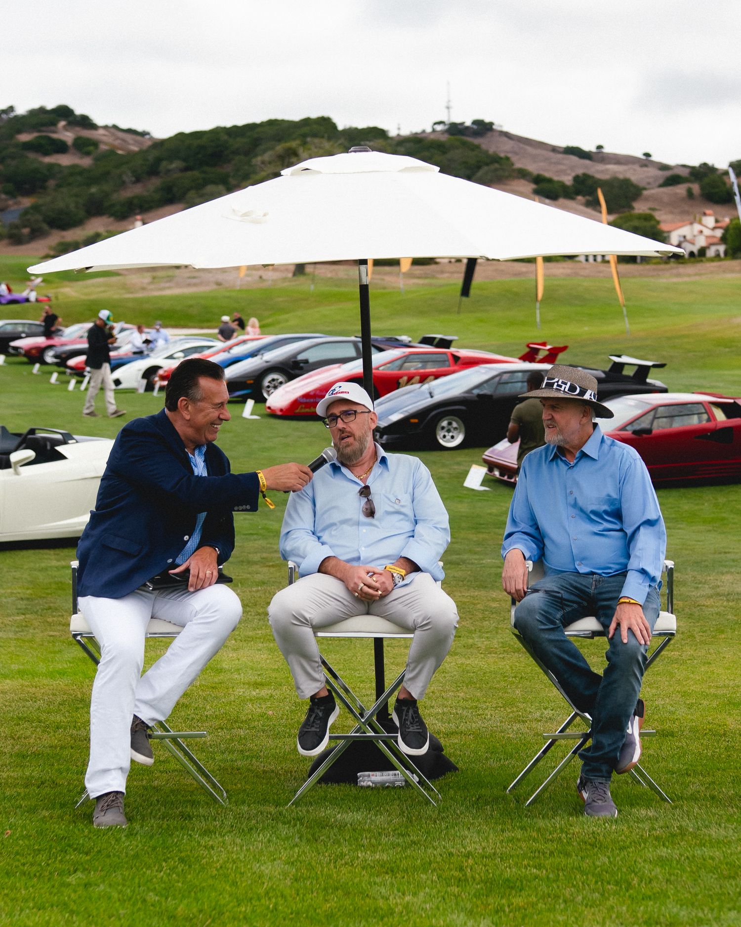 Three men conversing under a white umbrella at an outdoor classic car show on a green lawn.