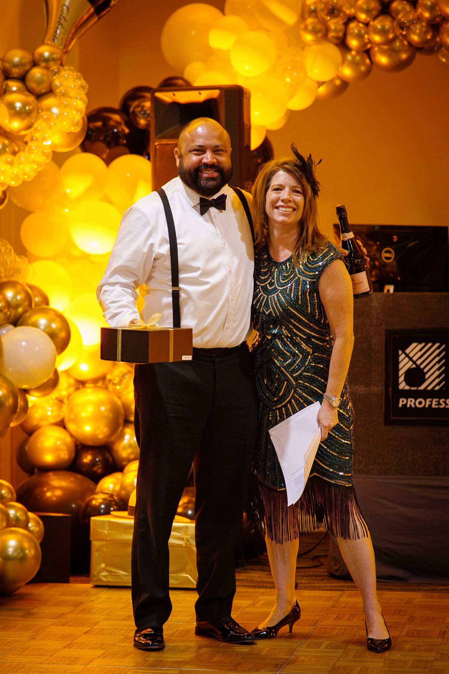 A man and woman pose together at a glamorous gold-themed gala event with balloon decorations.