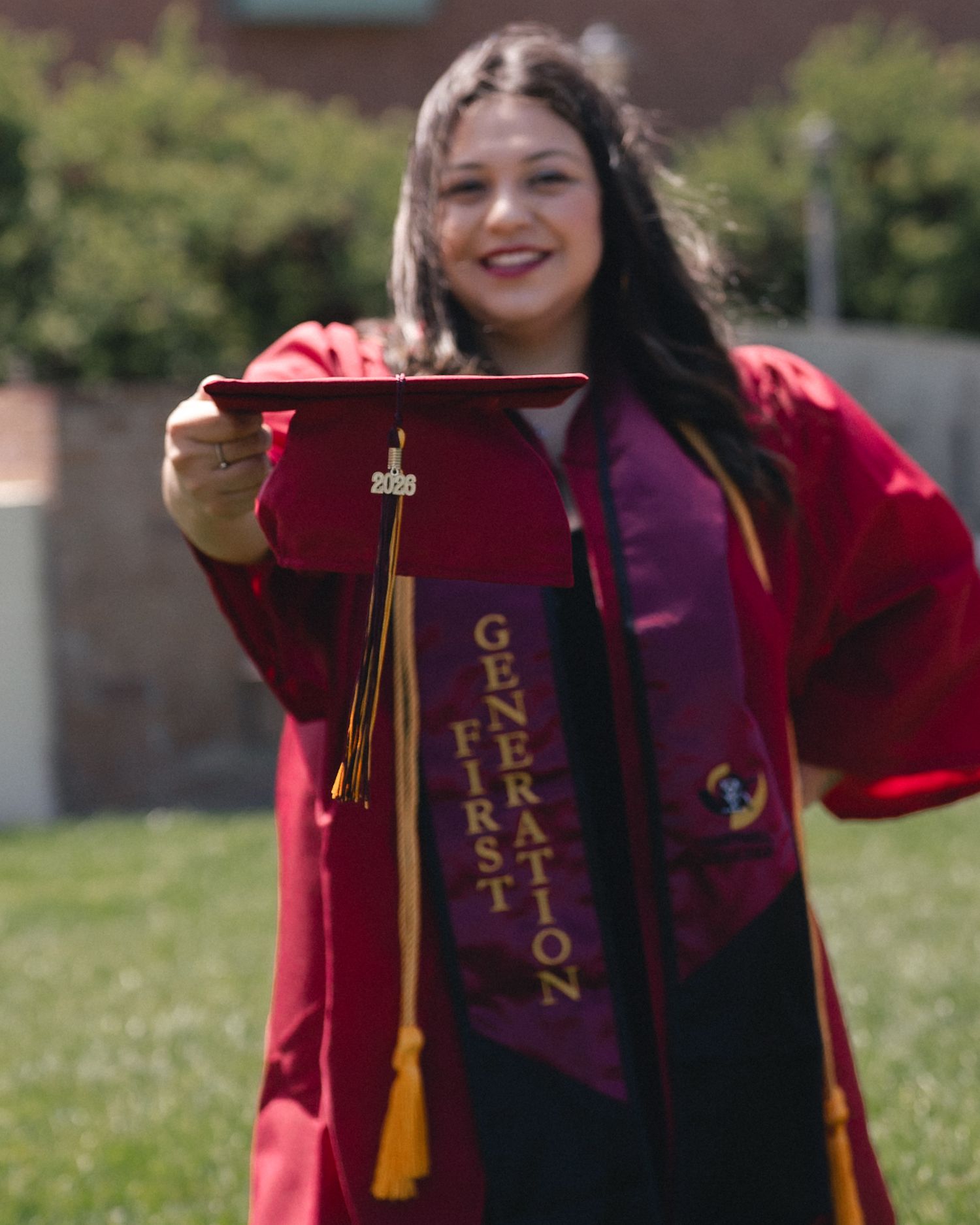 A smiling graduate in red regalia holds her cap, wearing a First Generation stole at her graduation ceremony.