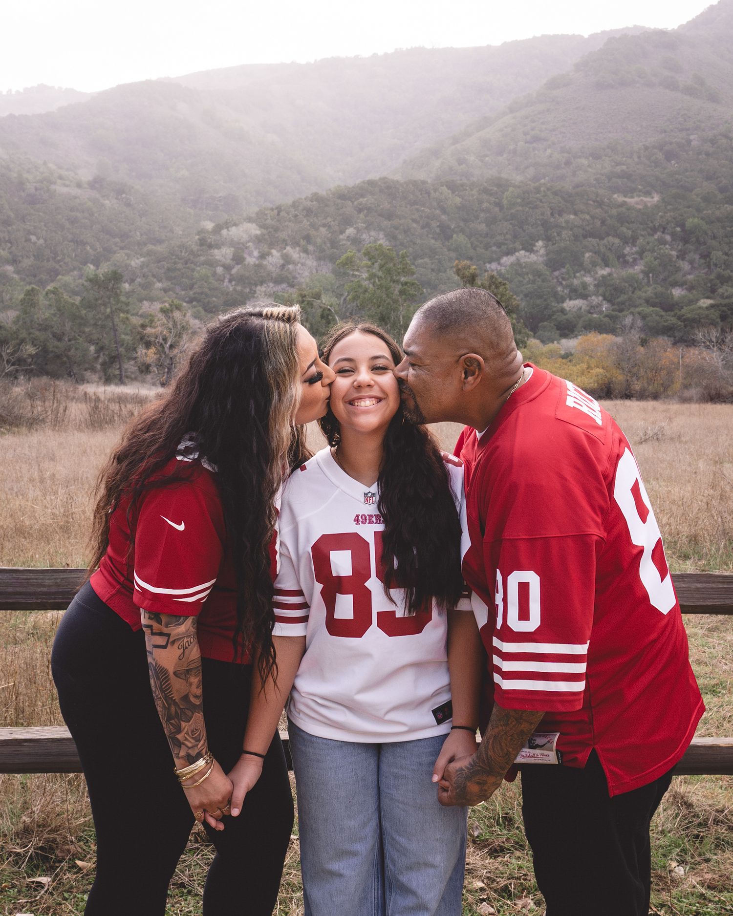 A family in San Francisco 49ers jerseys shares kisses outdoors, with green hills and a wooden fence in the background.