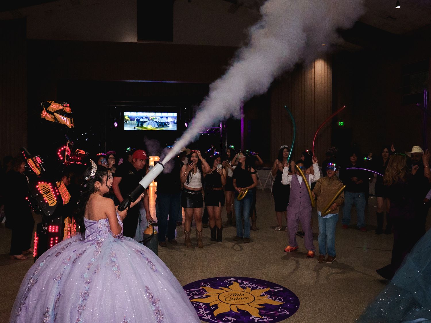 Quinceañera celebration with guests dancing, fog machine effect, and colorful lighting in a festive ballroom.