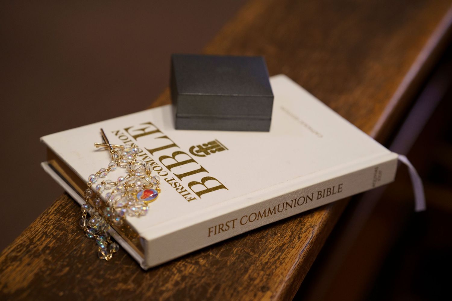 A white Bible and small black box rest on a wooden surface with a delicate silver cross necklace draped over them.