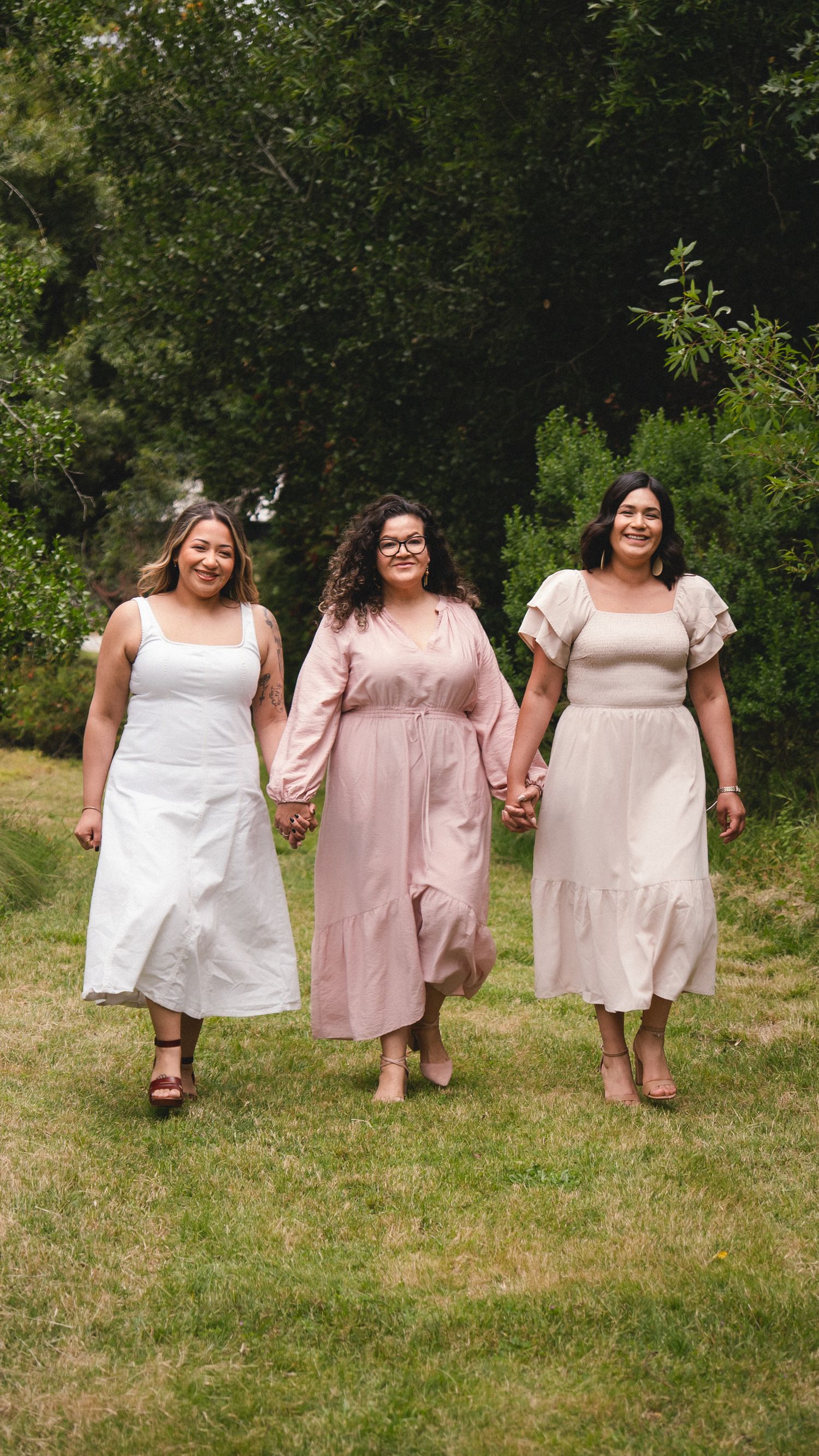 Three women in pastel dresses hold hands walking through a lush green park setting.